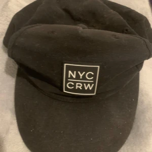 Svart keps med NYC CRW patch - Säljer en svart keps från NYC CRW med en tydlig vit patch framtill. Kepsen har klassisk form, böjd skärm och justerbar snapback baktill. Tillverkad i bomullstyg och passar perfekt till streetwear-stil.