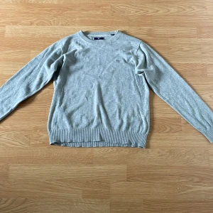 Ljusgrå stickad tröja från GANT - Säljer en klassisk ljusgrå stickad tröja från GANT. Tröjan har rund halsringning, ribbade muddar vid ärmslut och nederkant samt en liten broderad logga på bröstet. Perfekt för en stilren och enkel look.