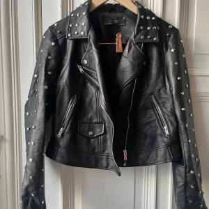 Svart skinnjacka med nitar från Zara - Snygg svart fejk skinnjacka från Zara med coola nitar på axlar och ärmar. Jackan har en unik broderad dödskalle på ryggen och dragkedjor på fickorna. Perfekt för att ge din outfit en edgy touch. Använd få tal gånger, inköpt för 6 år sedan, har blivit skrynklig efter ha varit i förrådet, liten i storlek och kort   Hund finns i hemmet, Skicka meddelande för mer info och bilder :)