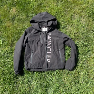 Svart vindjacka från Moncler - Säljer en svart vindjacka från Moncler med huva och dragkedja framtill. Jackan har Moncler-logga på ärmen och diskret logotyptryck längs dragkedjan. Perfekt för blåsiga dagar och snygg till streetwear. Passade inte mig riktigt. Pris kan diskuteras. Inte äkta