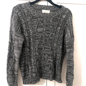 Gråmelerad stickad tröja - Säljer en gråmelerad stickad tröja med kabelstickat mönster. Tröjan har rund halsringning och långa ärmar, perfekt för kyligare dagar. 