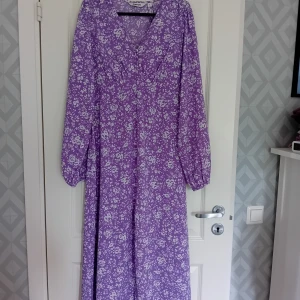 Långklänning  - Somrig långklänning i storlek S från Kappahl med fina detaljer. Lila med vita blommor, v-ringad med knappar fram. Helt ny och oanvänd med prislappen kvar. 200 kr. 