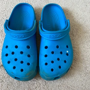 Blå Crocs sandaler - Säljer ett par klassiska blå Crocs sandaler med justerbar hälrem och ventilerande hål upptill. Tillverkade i lätt syntetmaterial som är bekvämt och vattentåligt. Perfekta för sommardagar eller som inneskor. Finns en fläck på, går med högst sannolikhet att ta bort.