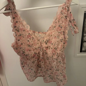 Blommig spetslinnetopp med rosett - Supersöt blommig linnetopp i ljusrosa med spetsdetaljer och knytband på axlarna. Toppen har volangkant och är ärmlös, perfekt för varma dagar eller lager på lager.