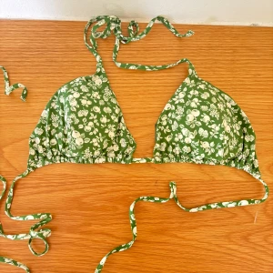 Grön blommig trekantsbikini - Säljer en grön trekantsbikini med vitt blommönster och knytband i nacken och ryggen. Perfekt för sommardagar på stranden. Bikinin har klassisk triangelmodell och tunna band.