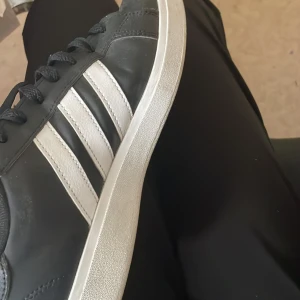 Svarta Adidas sneakers med vita ränder - Klassiska svarta Adidas sneakers med tre vita ränder på sidorna och vit sula. Skorna har snörning och rund tå, perfekt för en sportig och stilren look. Adidas-logga syns på plösen.