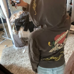 Ed Hardy hoodie  - Säljer en jättesnygg brun hoodie från Ed Hardy med dragkedja och broderad design med hjärta och text på ryggen och bröstet. Väldigt fin men inte min stil längre så kommer ej till användning! Ena dragkedjan är fast där nere men inte den övre så den går att dra upp och ner fortfarande 💓använd fåtal gånger💞Storlek M men sitter som S/M!