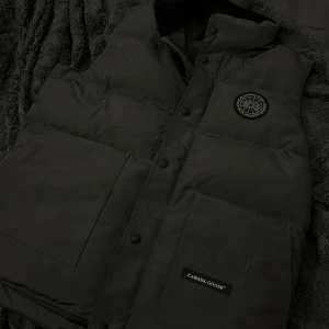 Svart dunväst från Canada Goose - Säljer en svart dunväst från Canada Goose med klassisk logotyp på bröstet och praktiska fickor framtill. Västen har knappar och hög krage, perfekt för lager-på-lager-stil under kyliga dagar.