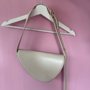 Beige axelväska i syntet - Säljer en stilren beige axelväska med justerbart axelband. Väskan har en halvmåneformad design och är tillverkad i slätt syntetmaterial. Perfekt för att bära det viktigaste och passar till många olika outfits.