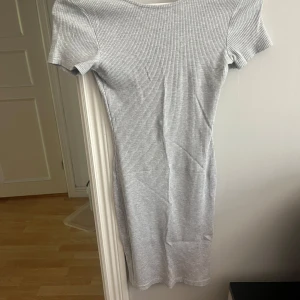 Ljusgrå ribbad kortklänning - Säljer en ljusgrå ribbad kortklänning med korta ärmar och djup, öppen rygg. Klänningen har en tight passform och är perfekt för sommardagar. Superenkel att styla med sneakers eller sandaler.