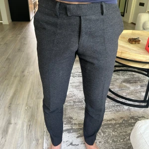 Mörkgrå kostymbyxor från Tiger of Sweden - Snygga mörkgrå kostymbyxor från Tiger of Sweden med normal passform och raka ben. Byxorna har pressveck, bälteshällor och diskreta bakfickor med knapp. Storlek: 48