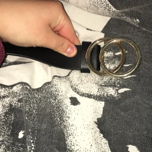 Svart bälte med dubbla guldringar - Säljer ett svart bälte med två stora, runda guldfärgade ringar som spänne. Bältet har en stilren och modern design, perfekt för att lyfta vilken outfit som helst. Materialet är slätt och bältet är brett.