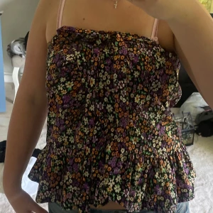 Blommig linnetopp med volang - Säljer en söt linnetopp med blommigt mönster i lila, orange, vit och grön. Toppen har smala axelband och volang nertill, perfekt för varma dagar. Från pull and bear 