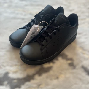 Svarta sneakers från Adidas - Stilrena svarta sneakers från Adidas med diskret perforerad logga på sidan och klassisk snörning. Skorna har en platt sula och är tillverkade i syntetmaterial. Perfekta för en clean och enkel look.