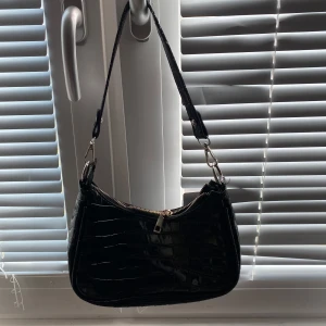 Svart axelväska med krokodilpräglat mönster - Säljer en svart axelväska i syntetmaterial med krokodilpräglat mönster. Väskan har en silvrig dragkedja upptill och justerbart axelband. Perfekt storlek för det viktigaste och snygg detalj till din outfit.