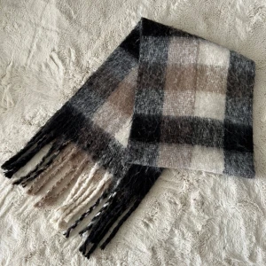 Rutig halsduk från Monki - Mysig rutig halsduk från Monki i svart, grått, beige och vitt. Halsduken har långa fransar i ändarna och är tillverkad i ett mjukt, fluffigt material som passar perfekt för kyliga dagar.