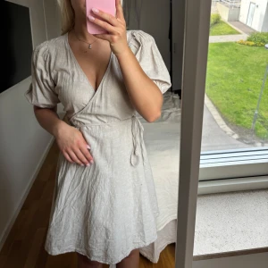 Beige omlottklänning i linne - Säljer en beige kort omlottklänning i linne med puffärmar och knyt i midjan. Klänningen har v-ringning och en lätt, luftig känsla – perfekt för varma dagar. Aldrig använd 
