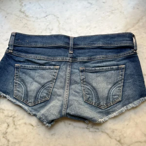 Blå jeansshorts från Hollister - Snygga blå jeansshorts från Hollister med fransig kant och klassiska bakfickor. Shortsen har låg midja och är i en klassisk denimtvätt med slitningar framtill. Perfekta för varma dagar.
