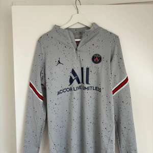 PSG långärmad träningströja från Jordan - Säljer en grå långärmad PSG träningströja från Jordan med mörkblå och röda detaljer på ärmarna. Tröjan har ett diskret prickigt mönster och dragkedja vid halsen. PSG-logga och sponsortryck på bröstet.
