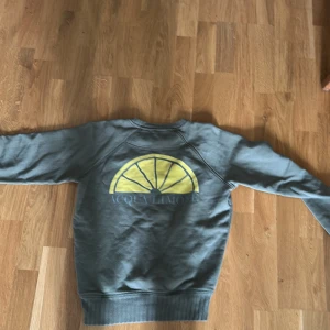 Grå sweatshirt från Acqua Limone - Säljer en grå sweatshirt från Acqua Limone med deras klassiska gula citrontryck på ryggen. Tröjan har rund halsringning och ribbade muddar vid ärmslut och nederkant. Perfekt för en avslappnad stil.