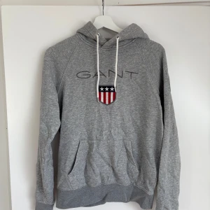 Grå hoodie från GANT - Säljer en klassisk grå hoodie från GANT med broderad logga och amerikanskt sköldmärke på bröstet. Tröjan har huva med vita snören och en stor magficka. Perfekt för en avslappnad stil.