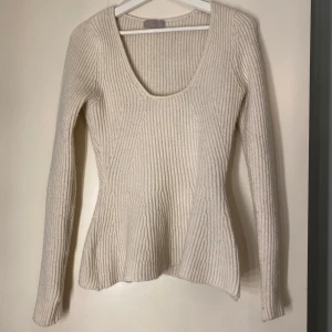 Beige/vit ribbad stickad tröja - Säljer en beige/vit stickad tröja med ribbad struktur och djup rundringning. Tröjan har lång ärm och en lätt utsvängd passform nedtill. Perfekt för lager-på-lager och enkel att styla till olika outfits.