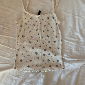 Vit blommig linnetopp från H&M Divided - Söt vit linnetopp från H&M Divided med små blå blommor och spetsdetaljer vid axelbanden. Toppen har en ribbad struktur och vågig kant nertill. Perfekt för varma dagar!