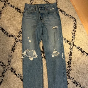 Levis 501 slitna jeans med hål - Klassiska Levis 501 jeans i ljusblå denim med slitningar och hål på benen. Medelhög midja och raka ben ger en tidlös look. Storlek 28/28.