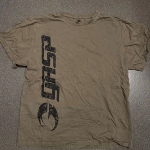 Olivgrön t-shirt från GASP - Säljer en olivgrön t-shirt från GASP med stort svart tryck både fram och bak. T-shirten har en klassisk passform och är kortärmad, perfekt för dig som gillar sportig stil.