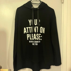 Svart hoodie med vit text - Säljer en svart hoodie med vit text framtill: 'YOUR ATTENTION PLEASE: NO ONE IS COMING TO SAVE YOU' och text på ena ärmen. Tröjan har huva med snören och långa ärmar. Perfekt för en avslappnad och cool stil.