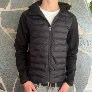 Svart pufferjacka från Polo Ralph Lauren - Säljer en svart cardigan från Polo Ralph Lauren med huva och dragkedja. Jackan har broderad logga på bröstet och är quiltad framtill med släta ärmar och rygg. Perfekt för kyliga dagar.