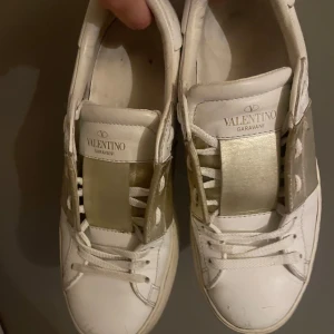 Valentino skor - Snygga vita sneakers från Valentino Garavani med bred guldfärgad rem över vristen och klassiska snördetaljer. Skorna har rund tå, platt sula och ikoniska nitar på hälen. Perfekta för dig som vill ha en stilren men lyxig look. Säljer på grund av att dom är lite för stora för att jag ska kunna ha dom bekvämt❤️ storlek 37 skulle säkert passa 37’5 vissa säger 38. Andvänt men riktigt fint skick. Skriv vid frågor och funderingar, priset kan diskuteras vid snabb affär!❤️