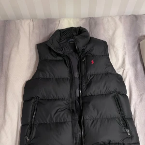 Svart dunväst från Polo Ralph Lauren - Säljer en svart dunväst från Polo Ralph Lauren med dragkedja och två sidofickor med dragkedja. Västen har en klassisk passform och är perfekt för lager-på-lager. Märkets logga i rött broderad på bröstet. Storlek M men passar även S nypris 3499kr