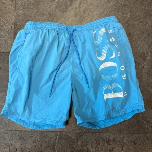 Blå badshorts från BOSS - Snygga blå badshorts från BOSS med vit logga längs ena benet. Shortsen har elastisk midja med blå snörning och innerbyxa i mesh med en bakficka och en innerficka. Perfekta för stranden eller poolen.