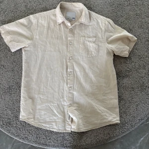 Beige kortärmad skjorta från 157 - Säljer en stilren beige skjorta från 157 i storlek M. Skjortan har korta ärmar, klassisk krage, bröstficka och knäppning framtill. Perfekt för varma dagar och enkel att matcha med olika outfits.