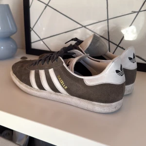 Adidas Gazelle grå/vita sneakers - Säljer ett par klassiska Adidas Gazelle sneakers i grå mocka pga jag inte använder dem längre. Skorna har svart snörning, men kan skickas med, med vita.sula. Storlek 44 2/3. Lite smuts på skorna men det går enkelt bort med lite tvätt.