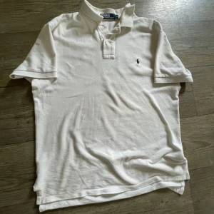 Vit pikétröja från Polo Ralph Lauren - Klassisk vit pikétröja från Polo Ralph Lauren med korta ärmar och knappar vid kragen. Tröjan har den ikoniska broderade loggan på bröstet och är tillverkad i bomull. Perfekt för en stilren och avslappnad look.