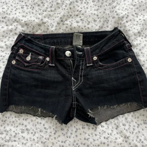 Svarta jeansshorts från True Religion - Säljer ett par svarta jeansshorts från True Religion med råa kanter och rosa kontrastsömmar. Shortsen har klassiska fem fickor och dekorativa bakfickor med broderad detalj. Perfekta för sommaren och har låg midja.