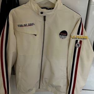 Vit racinginspirerad jacka Le Mans Steve McQueen - Vit jacka med racingstil, svarta och röda ränder på ärmarna och broderade detaljer. Märke och patchar på bröstet, dragkedja framtill och ståkrage. Fodret har ett tryck av Steve McQueen. Perfekt för motorsportfans. Använd endast någon gång. Inga fläckar eller skavanker. Bra passform sitter perfekt på mig och är 183cm. 