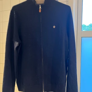 Mörkblå kofta från Morris - Säljer en stilren mörkblå kofta från Morris i 100% merinoull. Koftan har dragkedja framtill, ribbade muddar och en liten broderad logga på bröstet. Perfekt för lager-på-lager och passar till många olika outfits.