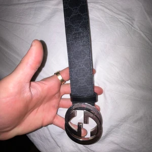 Svart skärp från Gucci med GG-spänne - Säljer ett svart skärp från Gucci med ett stort, runt GG-spänne i silverfärg. Skärpet är gjort i skinn och har diskret GG-mönster på framsidan. Perfekt accessoar för att lyfta din outfit.