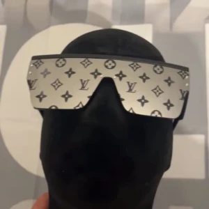 monogram solglasögon - Spektakulära solglasögon med spegelglas och ikoniskt monogrammönster över hela linsen. Rektangulär, oversized modell med silvrig finish och futuristisk vibe. Perfekt statement piece för dig som vill sticka ut.