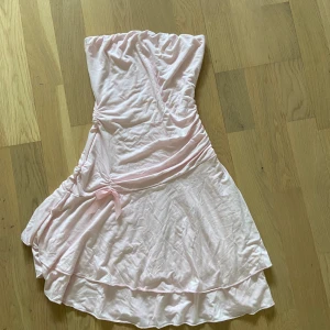 Ljusrosa axelbandslös omlottklänning - Söt ljusrosa axelbandslös klänning med omlottkänsla och volangdetalj nertill. Klänningen har en feminin passform och knyt i sidan för extra detalj. Perfekt för sommarens alla tillfällen!