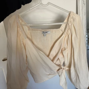 Beige omlottopp från Kappahl - Säljer en söt beige omlottopp från Kappahl med puffiga långärmar och smockad rygg. Endast använt en gång. Toppen har knyt i sidan och en lätt croppad passform. Perfekt för vår och sommar!