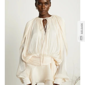 Champagnefärgad omlottklänning med volang - Säljer en champagnefärgad kortklänning med volangdetaljer och omlottknytning i midjan. Klänningen har långa, vida ärmar med resår vid handlederna och v-ringning med knytband. Tillverkad i ett lätt, glansigt material som ger en lyxig känsla. Säljer eftersom den var för stor på mig. Storlek M. Aldrig använd, prislappar finns kvar! klänningen är ifrån Miss Rosier