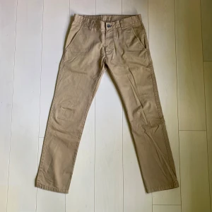 Beiga chinos från Dr Denim - Säljer ett par klassiska beiga chinos från Dr Denim med material av kvalitet. Byxorna har normal passform, raka ben och stängs med dragkedja och knapp. Tillverkade i 100% bomull och har fickor fram och bak. Perfekta för en stilren och avslappnad look.