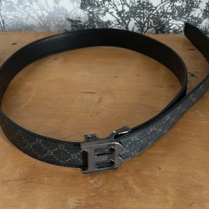 Svart skärp med B-spänne från Balmain - Säljer ett svart skärp från Balmain med ett stort B-format metallspänne i silver. Skärpet har diskret mönster i svart och grått längs hela bandet och är gjort i skinn. Perfekt accessoar för att lyfta din outfit.