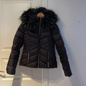 Svart pufferjacka med fuskpäls från Chiara Forthi - Säljer en svart pufferjacka från Chiara Forthi med stor huva och fuskpäls. Jackan har dragkedja framtill och två fickor med dragkedja. Snyggt mönstrad vaddering och figurnära passform. Perfekt för kyliga dagar!