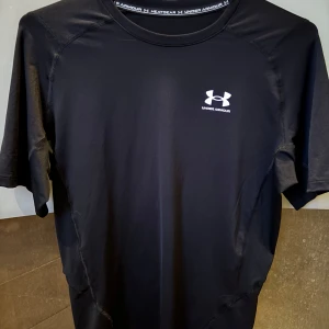 Svart kompressionströja från Under Armour - Säljer en svart kortärmad kompressionst-shirt från Under Armour HeatGear. Perfekt för träning eller sport, med logga på bröstet och stretchigt material som andas. Sitter tight och ger bra stöd under intensiva pass.