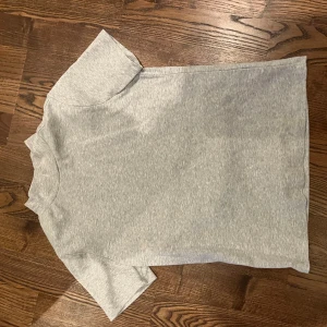 Grå basic t-shirt - En enkel och stilren grå t-shirt med lite högre hals och korta ärmar. Perfekt till jeans eller shorts för en avslappnad look. Gjord i mjukt bomullsmaterial som känns skönt mot huden.